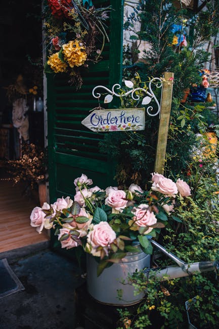 A lush floral display outside a floral shop features a bouquet of soft pink roses with velvety petals and green leaves arranged in a metal watering can, positioned on the ground near a green wooden sh