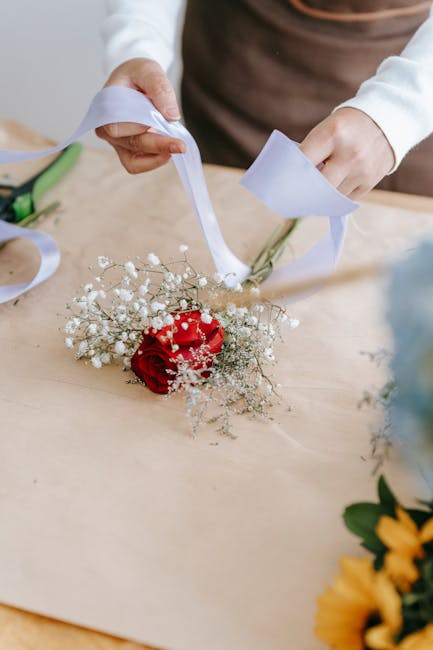 A person arranging a floral bouquet featuring a single vibrant red rose surrounded by delicate white baby's breath flowers. The bouquet is being wrapped with white ribbon, held over a light wooden tab