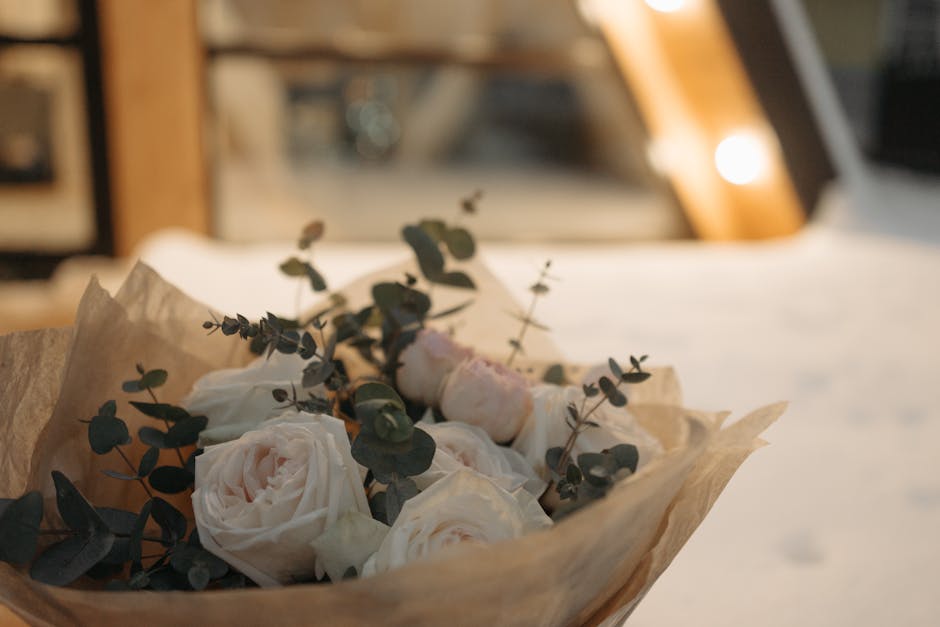 A bouquet of fresh, creamy white roses accented with sprigs of eucalyptus leaves, wrapped in light brown parchment paper. The roses have lush, velvety petals with delicate textures, arranged in a comp