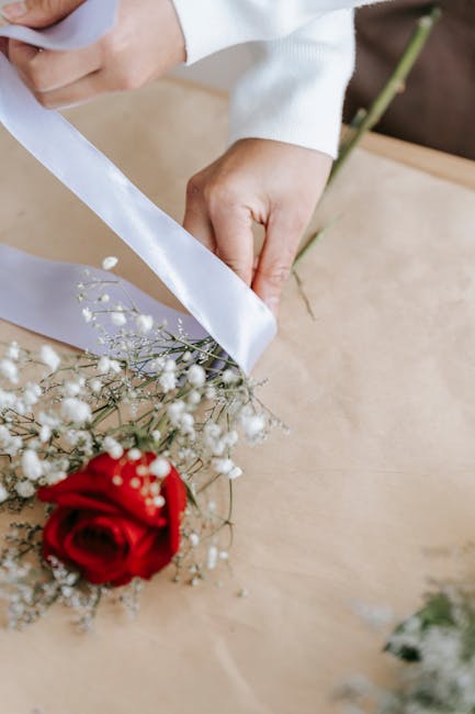 A florist's hands wrapping a fresh floral bouquet with white ribbon, featuring a single prominent red rose, delicate white baby's breath, and green foliage. The arrangement is being meticulously prepa