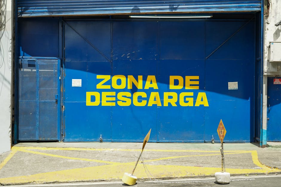 A large blue metal door with the words 'ZONA DE DESCARGA' painted in bold yellow letters, indicating a designated loading zone. Adjacent to the door, there is a small blue gate and two yellow traffic 