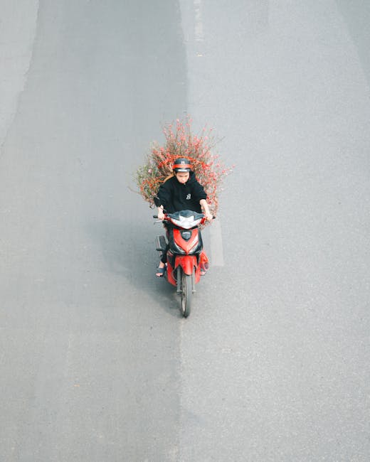 A woman riding a red scooter on a paved road, carrying a large, lush bouquet of mixed flowers with pink, red, and orange blooms, surrounded by green foliage, arranged in a natural, freeform style. The