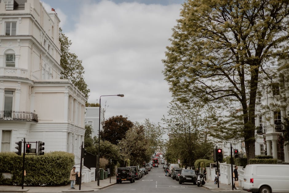 A residential street scene showing white Victorian-style buildings with decorative moldings and bay windows on either side. The street is lined with parked cars and bordered by lush green hedges and t