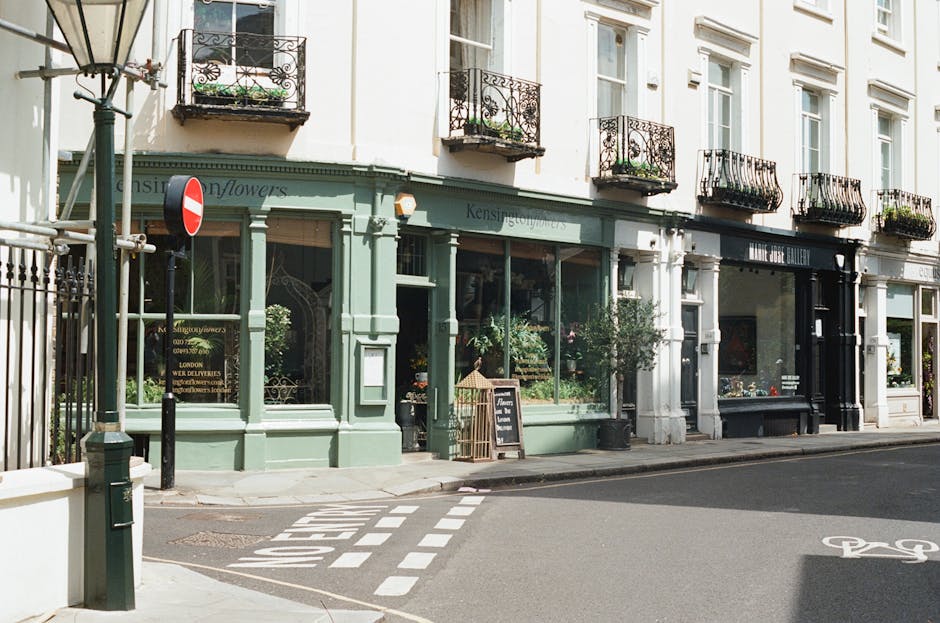 The image shows a charming flower shop with a soft mint-green facade and large glass windows displaying a variety of fresh flowers inside. The shop is situated on a busy street with white-painted road