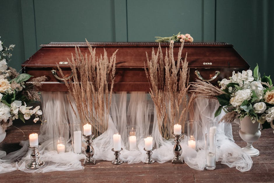 A funeral flower arrangement featuring white and cream roses, orchids, and greenery placed on either side of a polished wooden coffin with silver handles. Tall, dried pampas grass stems are positioned