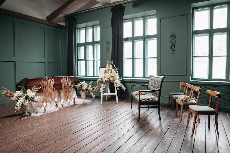 A floral tribute arrangement featuring white roses, lilies, and soft pastel blooms, complemented by dried pampas grass and foliage, set on an easel in a room with dark wooden flooring, teal-green pane