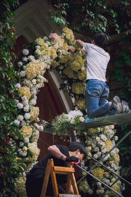 A young boy is kneeling on a scaffold, arranging a large floral installation composed of white and cream hydrangeas with some light pink blossoms, creating a decorative arch around a wooden door. The 