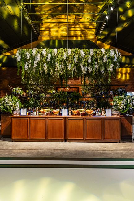 An elegant floral display features a large hanging arrangement of predominantly white flowers, including cascading wisteria or wisteria-like blossoms, suspended above a wooden bar counter. Below, two 