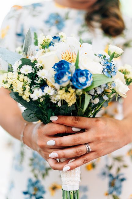 A bride holding a fresh flower bouquet arranged with white roses, blue lisianthus, small white daisies, and yellow-tinged flowers, wrapped in white lace and green foliage. The bouquet is held at waist