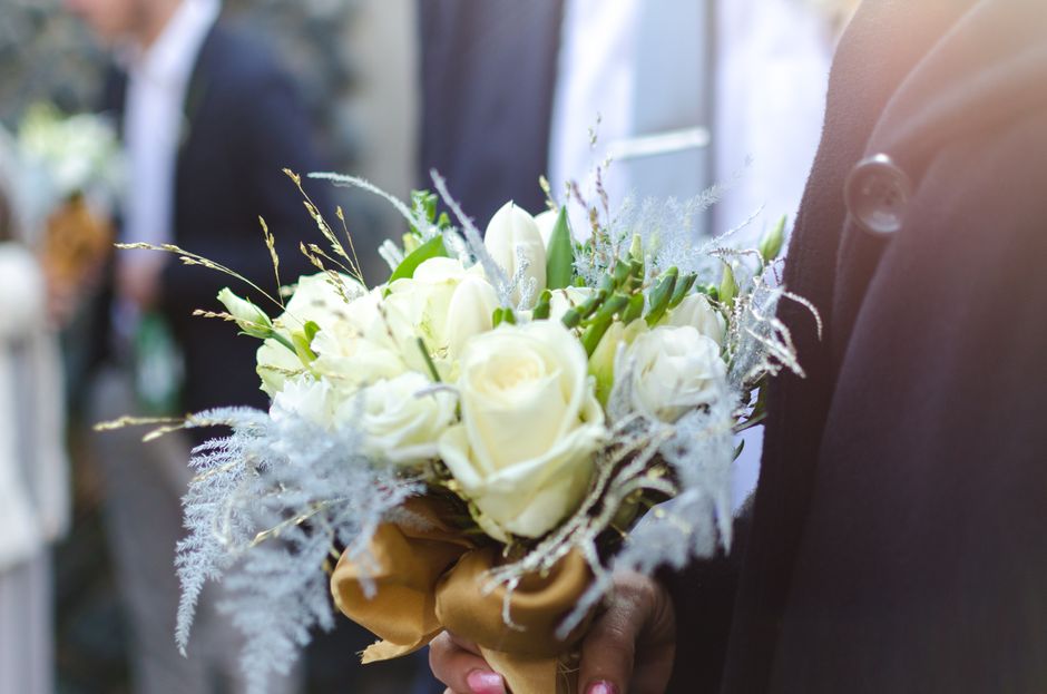 A floral arrangement featuring a bouquet of pristine white roses, interspersed with delicate greenery and wispy silver foliage, held by a person dressed in formal dark attire. The bouquet is wrapped i