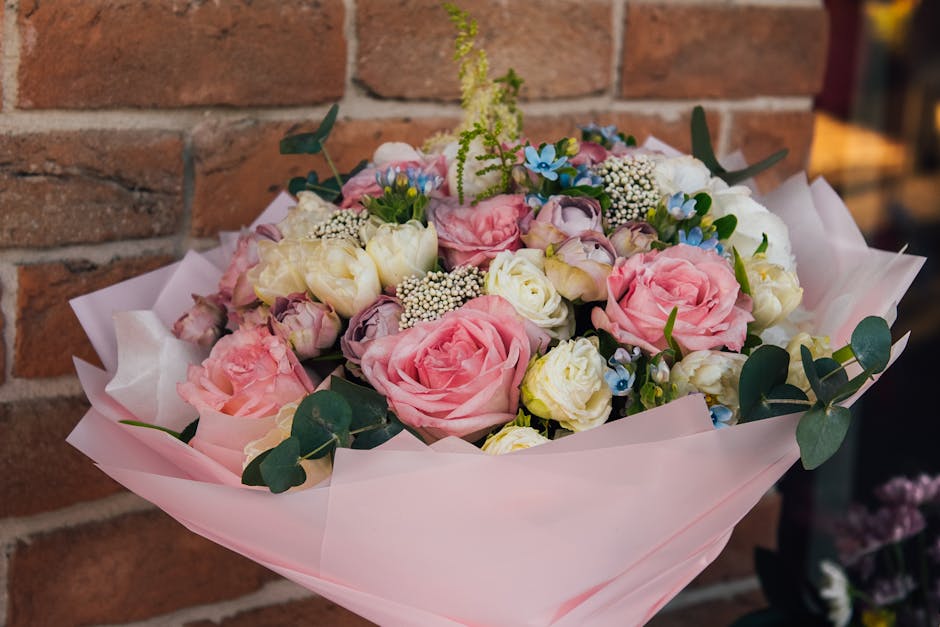 A floral bouquet featuring large pink roses, white lisianthus, and small blue forget-me-nots, accented with sprigs of white baby's breath and green eucalyptus leaves. The arrangement is wrapped in sof