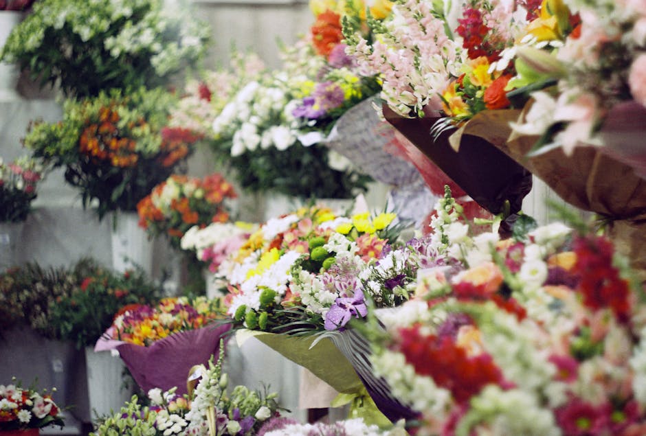A vibrant display of fresh floral bouquets arranged on a wall-mounted shelf, featuring a variety of flower types including lilies, roses, gerbera daisies, and stock. The bouquets showcase a wide palet