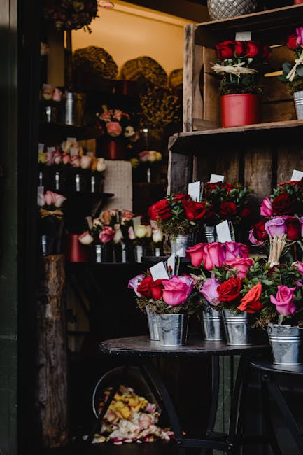 A display of fresh flower bouquets arranged in small metal buckets on a round black table inside a floral shop. The bouquets predominantly feature pink and red roses, complemented by smaller white and