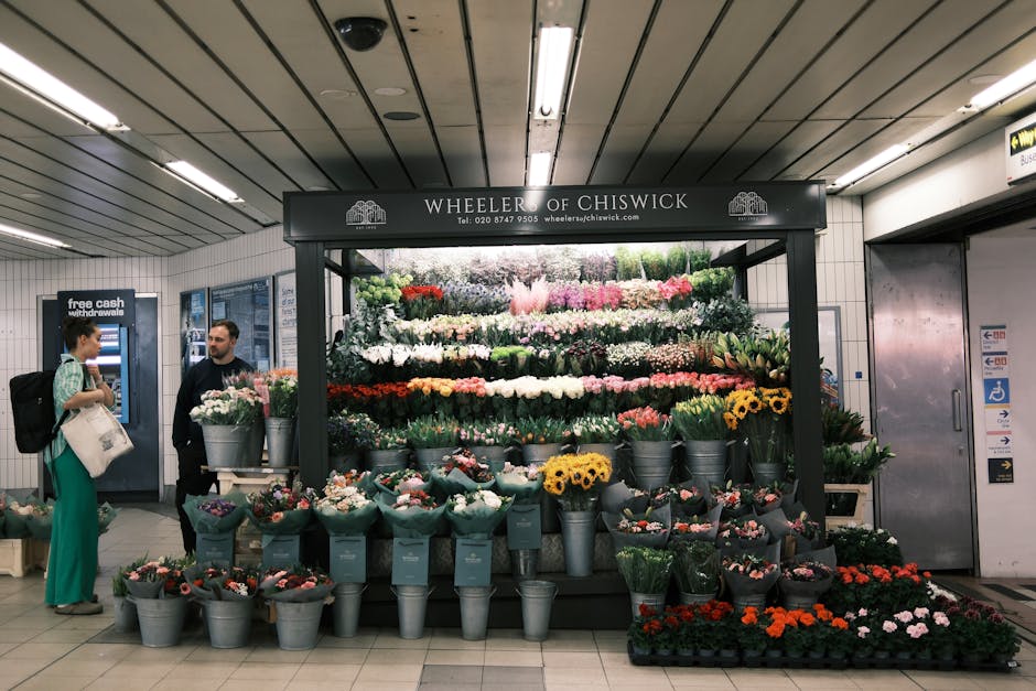 A flower stall named Wheeler of Chiswick is situated in an underground station, displaying an extensive array of fresh flowers arranged in buckets and on tiered shelves. The display includes vibrant b