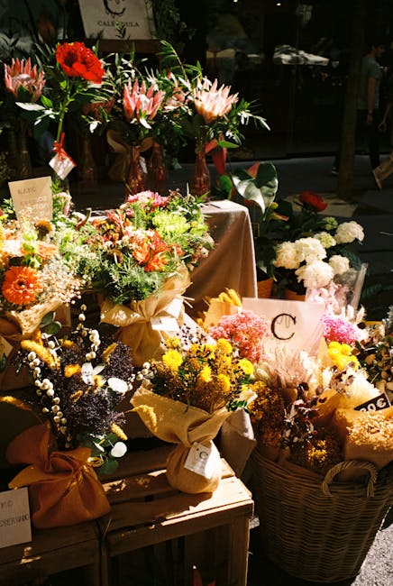 An assortment of vibrant floral arrangements displayed on a wooden table and in a wicker basket, featuring a variety of fresh flowers including pink lilies, red roses, white hydrangeas, yellow daisies